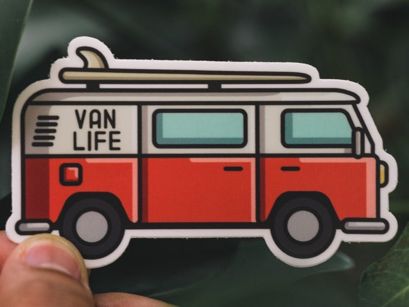 A hand holding a die-cut sticker of a red camper van with a surfboard.